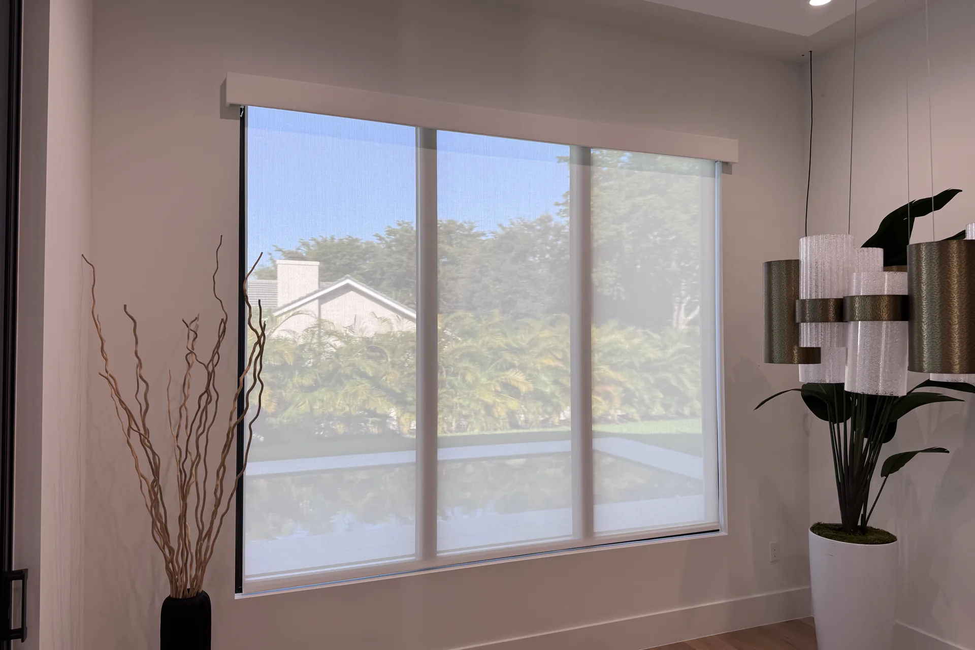 A large window with a white sheer roller shade in a white cornice, a pool and garden visible through the fabric