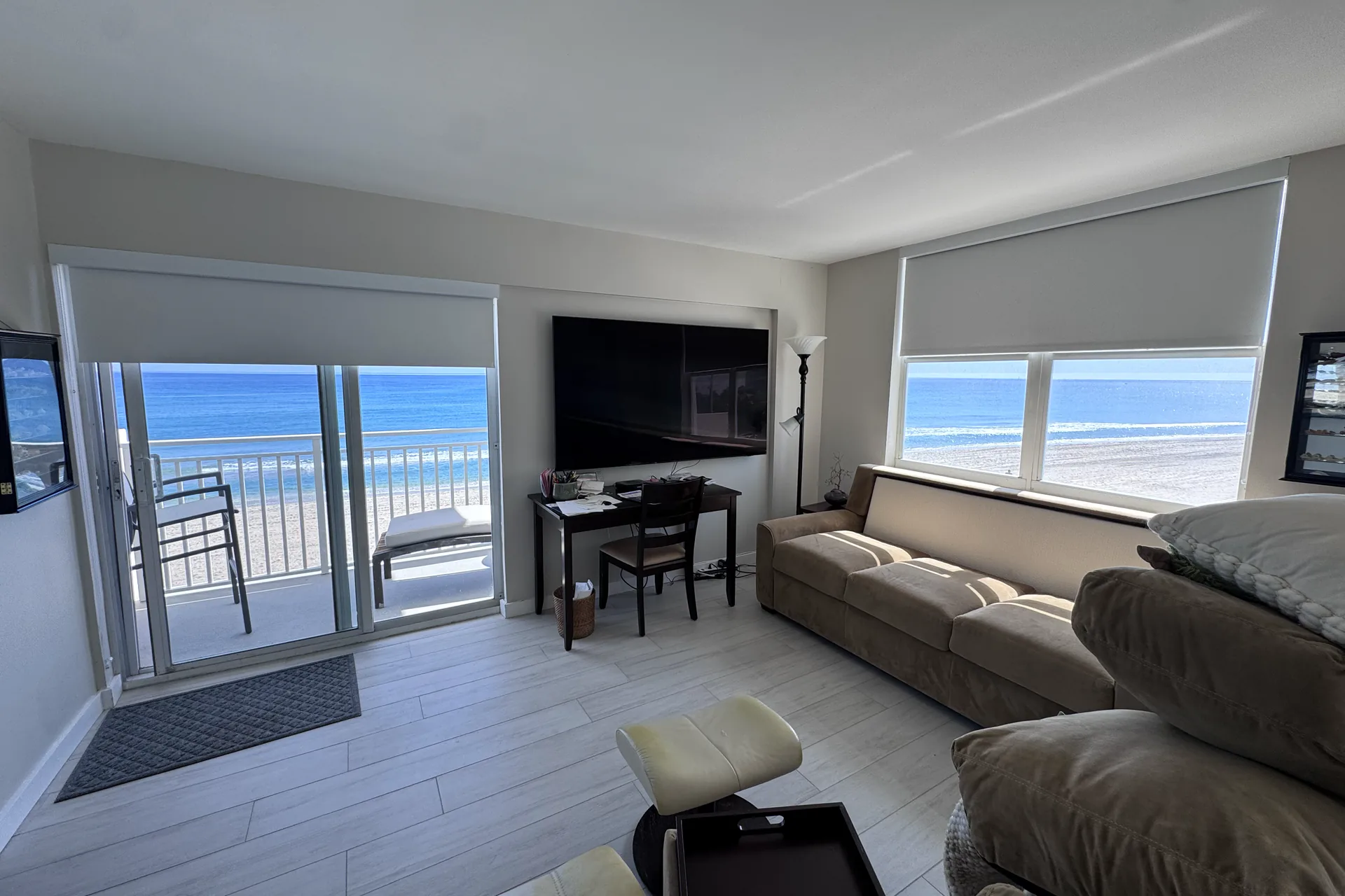 An oceanfront living room with white roller shades on a sliding door and a window, the beach beyond