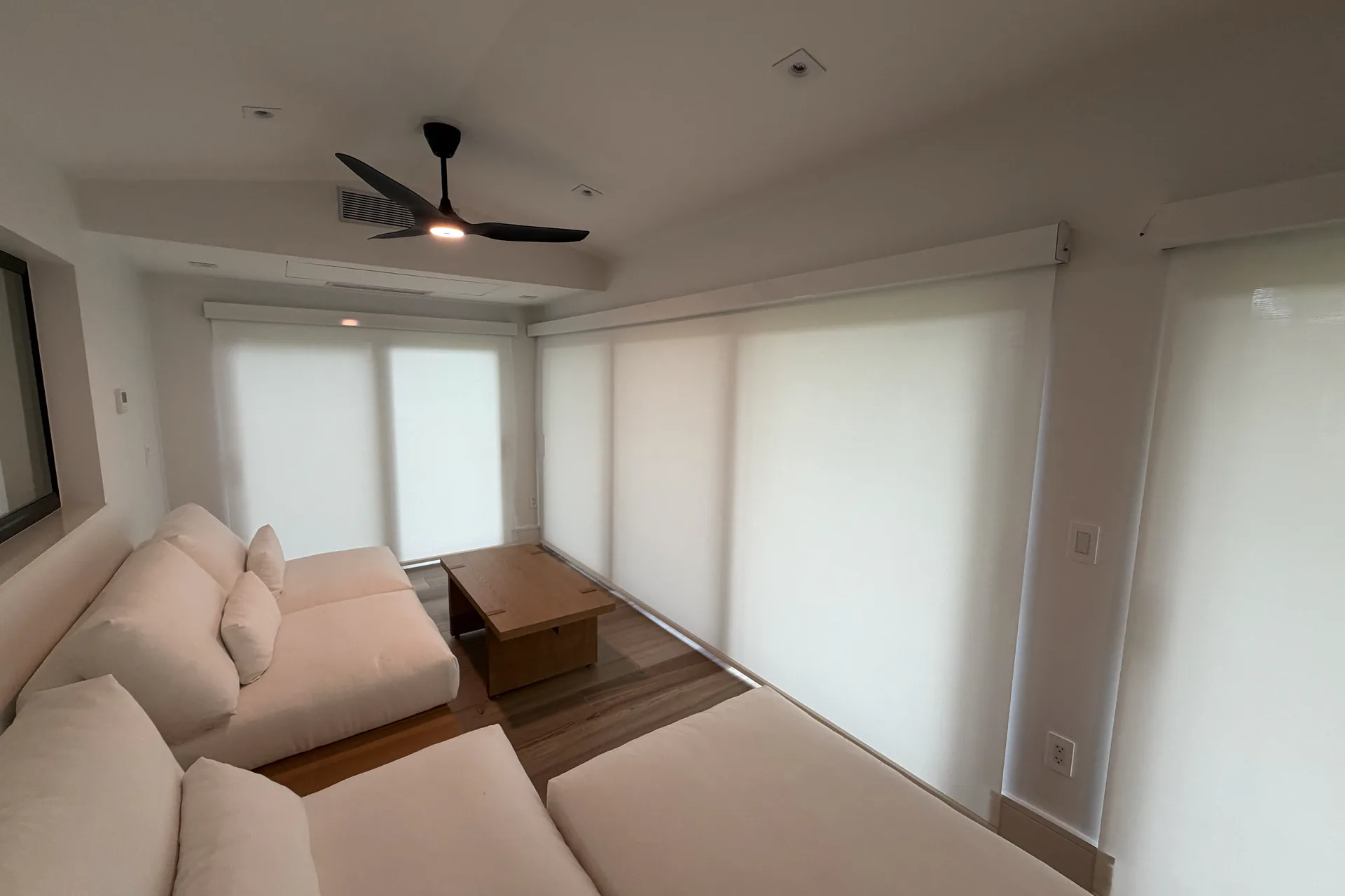 A room with large white roller shades on every window wall, a ceiling fan above a low sofa