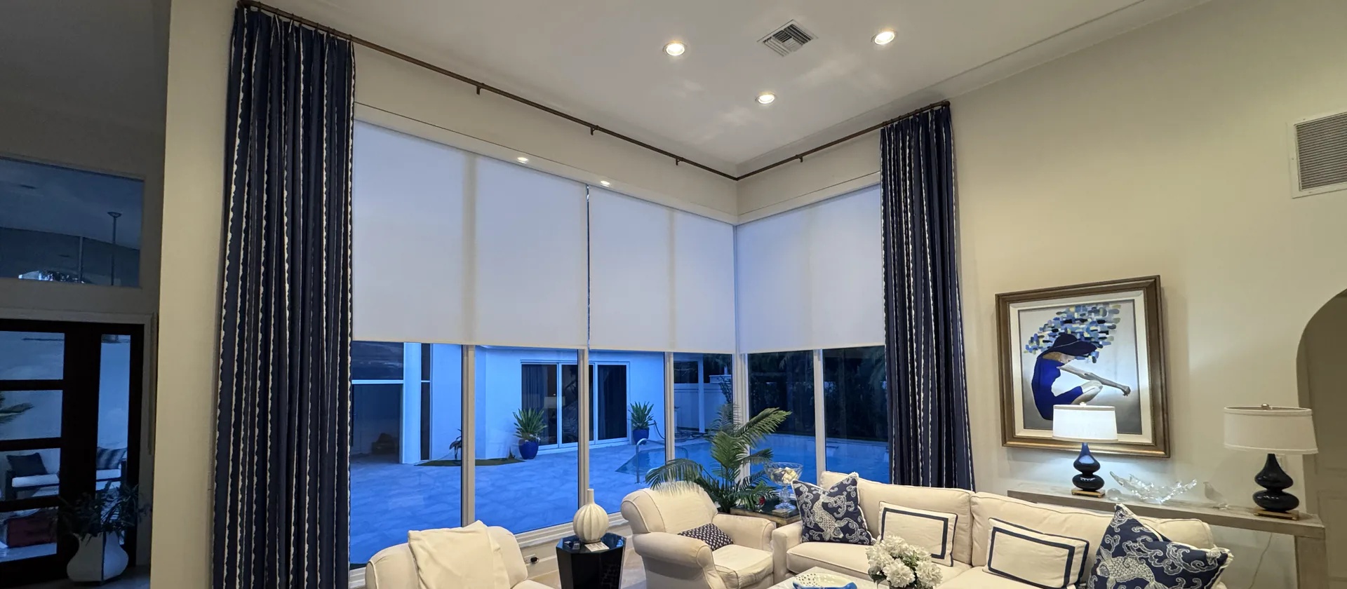 A tall living room at dusk with white roller shades on floor-to-ceiling windows and navy drapery on a rod, a pool courtyard beyond
